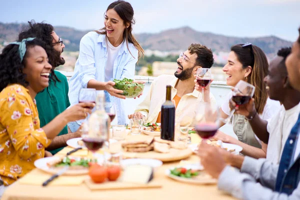 Genç, gülümseyen, beyaz bir kadın çatıdaki mutlu arkadaşlarıyla kutlamada sofraya yemek koyuyor. Bir grup neşeli çok ırklı arkadaş açık terasta barbekü partisi için toplandı..
