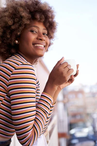 縦の写真 アフロの髪をした幸せな若い黒人女性がカメラの笑顔を見てポーズ 陽気なアフリカ系アメリカ人の少女が屋外でコーヒーを飲んでいます 肯定的な表現を持つ女性の肖像 — ストック写真
