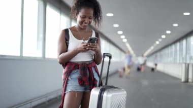 Genç Afrikalı Amerikalı kadın yaz tatili için uçak bileti aramak için havaalanında cep telefonunu kullanıyor. İnsanlar ellerinde bavullarıyla terminaldeki seyahat noktalarını kontrol ediyorlar..