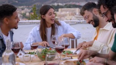 Çatıdaki barbekü partisinde etnik çeşitlilikte gençlerin gülümsemesi. Dışarıdaki partiyi kutlayan mutlu bir arkadaş grubu. Erkekler ve kadınlar yaz akşamları neşeyle yer ve içerler..