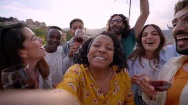 Neşeli, Afro-Amerikalı genç bir kadının çatıda arkadaşlarıyla parti hakkında blog için video çekmesi. İnsanlar kameraya bakıyor, dans ediyor ve şarap kadehleri tutuyor. Sosyal ağ.
