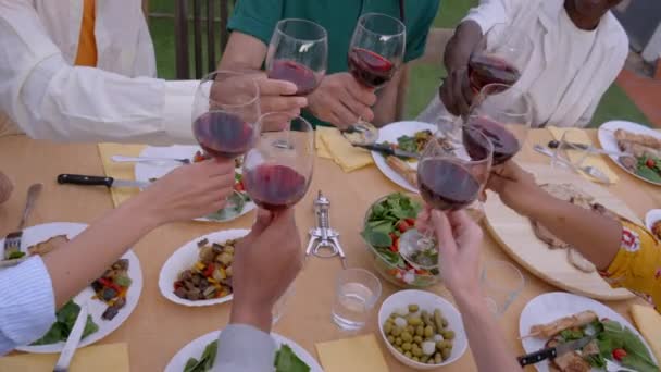 将盛满食物的红葡萄酒放在桌子上 手拉手敬酒 一群默默无闻的年轻朋友聚在一起庆祝烧烤派对3 人们坐在桌旁 欣赏着外面美丽的一天 — 图库视频影像