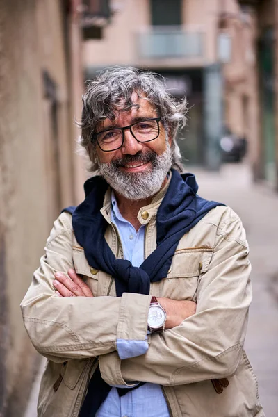 Dikey, gri saçlı, çapraz kollu, gülümseyen, mutlu bir adam. Şehir sokağındaki kameraya bakıyor. Grizli, olgun, neşeli ve açık hava portresi için kendinden emin bir beyefendi. Emekli insanlar ve kopyalama alanı
