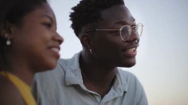 Kapalı alanda eğlenen genç, gülümseyen siyah çifte yakın çekim. Neşeli Afrikalı Amerikalı erkek ve kadın gün batımında sarılıp sohbet ediyorlar. İnsanlar romantik anları paylaşıyor. Kız ufku gösterir.