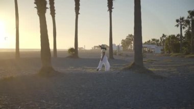 Altın gün batımında plajda yürüyen şapkalı Afrikalı genç bir kadının yavaş çekim görüntüsü. Yaz tatilinde gezen kadın turistin silueti. İnsanlar güzel bir yerde gezip eğleniyorlar.. 