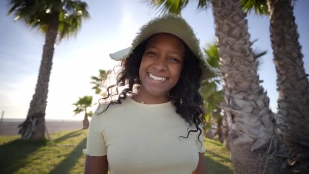 アフリカ系アメリカ人の女性がゆっくりとビーチのカメラで笑っている 若い女の子は積極的な態度で夏休みに屋外でポーズをとります 陽気世代背景に美しい夕日とZの人々 — ストック動画