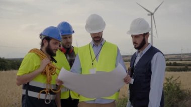 Bir grup beyaz mühendis kağıt planları kullanarak rüzgar türbinlerini kontrol ediyor. Yenilenebilir enerji teknolojisi ve sürdürülebilirlik. Gelecek ve çevre bakımı için alternatif çözümler.