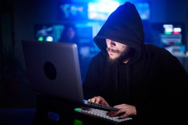 Tişörtlü ve kapüşonlu gizli bilgisayar korsanı güvenlik sistemlerini hackliyor. Karanlık odada mavi neon ışıklarla oturan tanınmayan sakallı adam. Siber saldırı, kötü amaçlı yazılım ve siber güvenlik kavramı.