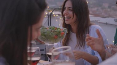 Beyaz bir kadın yemek yerken arkadaşlarıyla gülüşüyor. İnsanlar dışarıda barbekü kutlaması için toplandı. Yavaş çekim mutlu grup arka planda şehrin çatısında yemek yiyor.. 