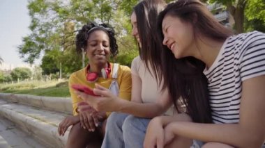 Dışarıda parkta cep telefonu kullanan bir grup çok ırklı mutlu genç kadın. Güneşli yaz gününde cep telefonuyla eğlenen üç gülen arkadaş. Z nesli, flört uygulamaları ve sosyal ağlar.