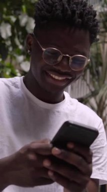 Üniversite kampüsünde cep telefonu kullanan ve kameraya bakan mutlu bir zencinin dikey videosu. Gülümseyen Afrikalı Amerikalı çocuk sosyal medyaya mesaj atıyor ve kontrol ediyor. Ağlara bağlı neşeli bin yıl.