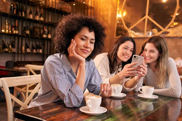 stock image Portrait of a happy Latin woman in a cafe bar looking at the camera. People indoors staring at a smartphone at the same table while having coffee. Female girlfriends spending time together