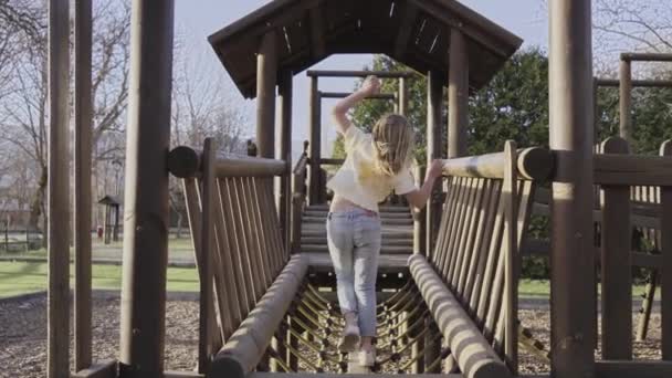 활동적 소녀는 놀이터 다리에서 있습니다 금발의 소녀는 나무로 집에서 카메라를 — 비디오