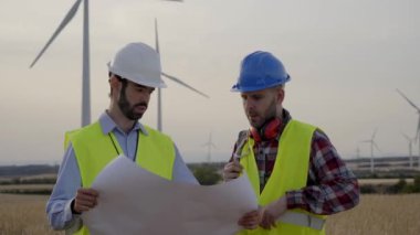 İş kıyafetli iki rüzgar mühendisi sahada bir rüzgar türbini sistemi kurma planlarını tartışıyor ve inceliyor. Profesyonel teknisyenler bir rüzgar santralinin önünde toplandı. Temiz yenilenebilir enerji kavramı.