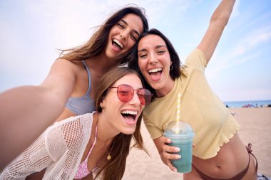 Sahilde selfie çeken, yaz kıyafetleri içinde üç heyecanlı beyaz arkadaş. Bir grup gülen kadın tatilin tadını çıkarıyor. Neslinin neşeli güzel kızları cep telefonlu fotoğraf için poz veriyor.