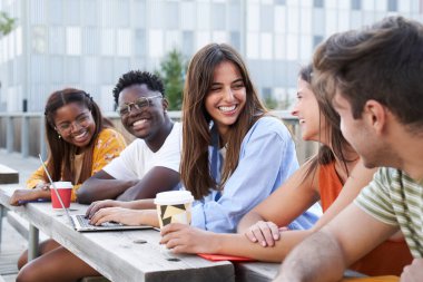 Üniversite kampüsünde oturmuş gülümseyen bir grup öğrenci. Bir kız dizüstü bilgisayar kullanıyor ve diğerleri kahve içiyor. Çeşitli ülkelerden oluşan çok ırklı bir gruptur..