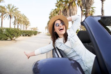 Genç, güzel, beyaz bir kadın, Cabrio arabasında eğleniyor. Güneş gözlüğü takıyor ve çok gülüyor. Arka planda üstü açık bir arabada giderken palmiye ağaçları var. Tatiller ve özgürlük kavramı