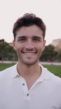Parkta kameraya bakan çekici, gülümseyen bir gencin dikey portresi. Beyaz erkek yakışıklı model dışarıda fotoğraf çektirmek için neşeli pozlar veriyor. Odaklanmamış arkaplan kopyalama alanı. Pozitif ifade 