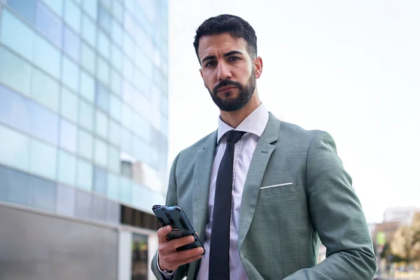 Portrait of young handsome confident business man using cell phone while taking break outdoors. Formal professional standing with mobile in hand and looking at camera serious expression, copy space.