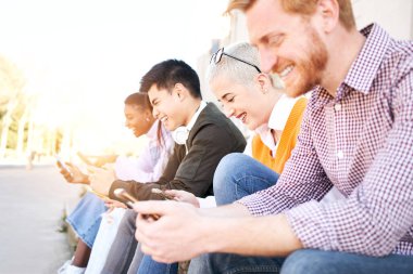 Teknoloji Bağımlısı gülümseyen bir grup öğrenci cep telefonu kullanıyor. Telefonlarındaki multimedyayı kontrol ediyorlar ve birbirlerine bakmıyorlar. Üniversite kampüsünün dışında gülüyorlar.