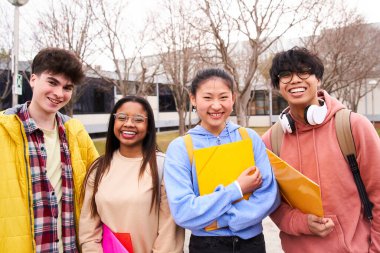 Ayakta durup kameraya bakan dört öğrencinin portresi. Üniversitenin dışında gülerler ve klasörleri tutarlar. Gen z