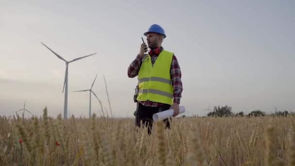 風力発電所に立っているウォルキー トーキーについて話す技術者 持続可能な再生可能エネルギーの熟練労働者 電力発電所について 屋外の仕事の制服の専門の技術者 — ストック動画