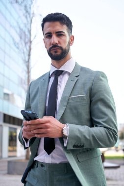 Dikey portre genç yakışıklı iş adamı cep telefonunu açık havada kullanıyor. Resmi profesyonel duruş, cep telefonu elinde ve kameraya bakıyor. Ciddi ifade, boşluğu kopyala. 