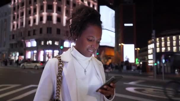 年轻的拉丁女性站在市中心 脸上挂着智能手机的笑容 灯光主街背景 复制空间 快乐的非洲女性在户外咨询和检查移动应用程序 — 图库视频影像