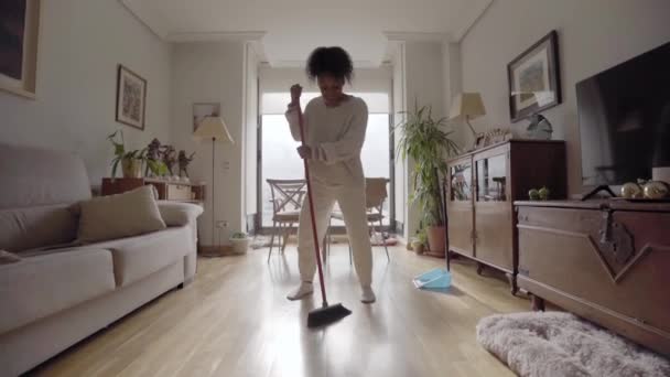 年轻的非洲裔美国女孩带着扫帚在客厅里跳舞唱歌 黑人用刷子和砂锅扫地来打扫房子 快乐的女人做家庭生活 — 图库视频影像