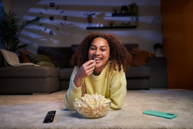 Afro Latin kadın kameraya bakıp oturma odasındaki halıya uzanmış patlamış mısır yiyor. Genç ve mutlu bir bayan gece bir kase yemekle sinemanın tadını çıkarıyor. Z jenerasyonu ve boş vakitler..