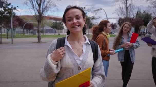 陽気な大学の慎重な女の子は外のカメラを見ている笑顔を浮かべます バックグラウンドで幸せなクラスメートとノートのフォルダを保持するヤングミレニアルの楽しい学生 試験期間中のキャンパスでの生活 — ストック動画