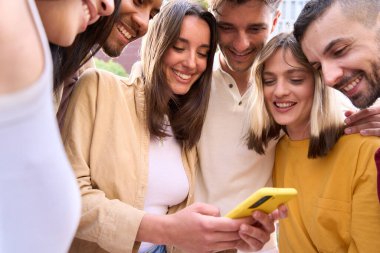 Genç bir grup insan dışarıda sosyal medyayı izlerken cep telefonuyla eğleniyor. Akıllı telefon uygulamasındaki içerikten hoşlanan neşeli öğrenci topluluğu. Genç çok ırklı arkadaş grubu 