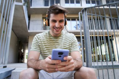 Kulaklık ve cep telefonu kullanan genç bir adamın portresi. Hücreye bakıyor ve gülümsüyor. Açık havada üniversitede oturuyor..