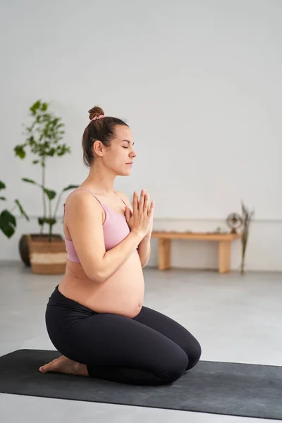 Vertical side view portrait young pregnant Caucasian woman doing yoga exercises meditating in class. Female fitness and future motherhood. Concept health care, mindfulness, relaxation and wellness.
