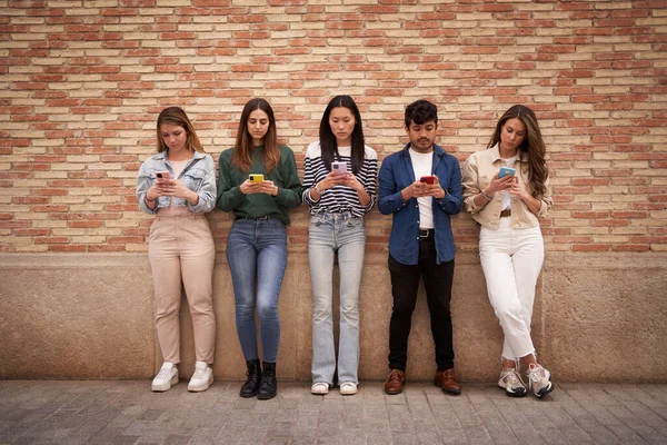 Multiracial serious group young generation z leaning on brick wall using phones outdoors. Worried student friends looking at mobile social media content. Boring people and technology addiction.