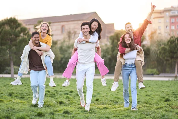 Group of funny pairs of friends piggybacking in park on sunny spring day. Three couples of cheerful multiracial young having fun outdoors. Community friendships between men and women in youth.