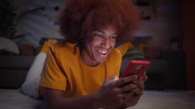 Oturma odasındaki halıya uzanmış akıllı telefon kullanan neşeli afro çocuğu yakından çek. Geceleri gülümseyen adam cep telefonunda sohbet ediyor. Bekar insanlar, randevu uygulamaları ve Z jenerasyonundaki sosyal ağlar..