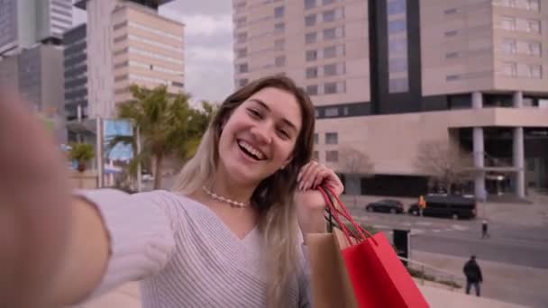 ショッピングで紙袋を持ち 外のモールに立っているカメラと話す陽気な若い女性ショップアホリックのビデオコール カメラに彼女の購入を示す割引販売でハッピーガール — ストック動画