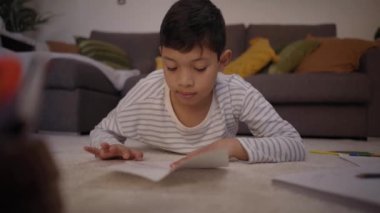 Latin çocuk evde yerde yatarak origami yapmaya konsantre oluyor. Rahat oturma odasındaki çocuk boş zamanlarında kağıt uçak yapıyor. Evde pozitif çocukluk hobileri kavramı.
