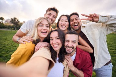 Bir grup neşeli çoklu etnik arkadaş dışarıda cep telefonlarıyla selfie çekiyorlar. Mutlu insanlar birlikte gülümsüyor ve parkta boş vakitlerin tadını çıkarıyor. Toplumda pozitif ve eğlenceli gençlik ilişkileri.