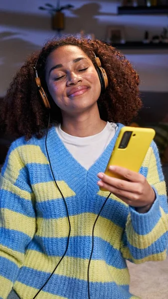 Vertical Young African American girl sitting in living room relaxing listening to music with headphones. Afro latina woman on carpet enjoying mobile phone music at home at night. Free time and leisure