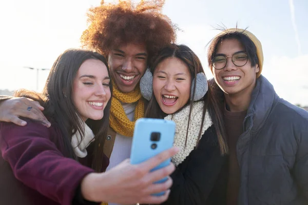 Group of excited friends enjoying taking selfie mobile photo looking at screen phone in warm clothes. Young smiling multiracial people having fun in cold and sunny winter day outdoor. Generation z.