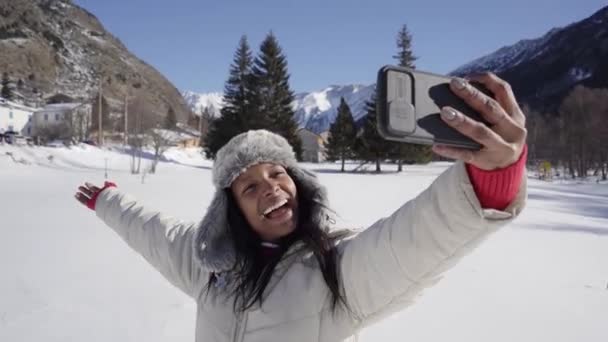 雪山で冬の日にセルフィーを取るアフリカ系アメリカ人の少女 冷たい屋外で写真のために面白い舌を出す女性 携帯電話を使用して暖かい服で美しい女性を笑う — ストック動画