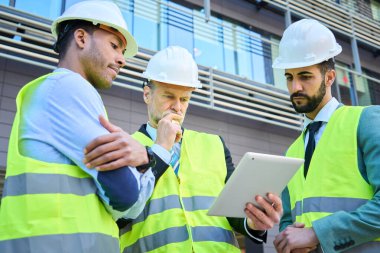 Birkaç mühendis dijital ekran kullanarak tablet ekrana odaklanmış görünüyor. Başarılı mimarlar yeni proje için toplanıyor. İnsanlar bir araya gelerek güvenlik kıyafetlerini tartıştılar.