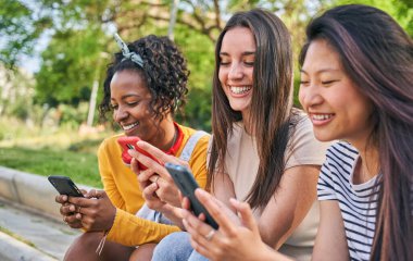 Dışarıda cep telefonu kullanan üç gülümseyen genç kadın. Bir grup mutlu arkadaş cep telefonundan eğlenceli sosyal ağları kontrol ediyor. Neşeli sınıf arkadaşları flört uygulamalarında dedikodu yaparlar. İnsanlar ve yeni teknolojiler.