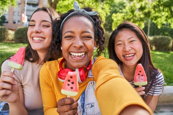 Selfie of three smiling young multi-ethnic women outdoors eating ice cream. Friends having a fun outdoors and enjoying the summer atmosphere together. Vacation time. Concept of friendship.