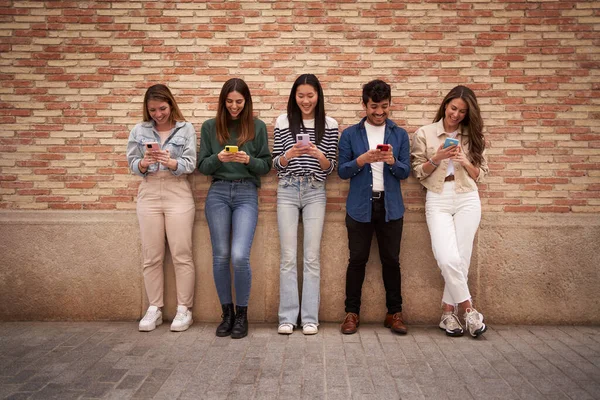 Multiracial smiling group young generation z leaning on brick wall using phones outdoors. Cheerful student friends looking at mobile enjoying social media content. Happy people, technology addiction.