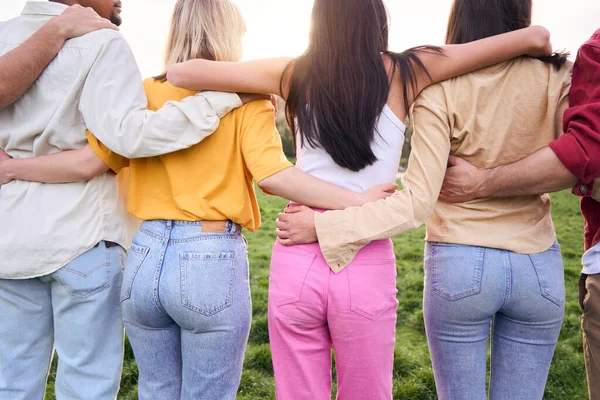 Multicultural group as team and community side by side in nature. Unrecognizable friends together in park hugging each other. Rear view photo of young people outdoors united in park.