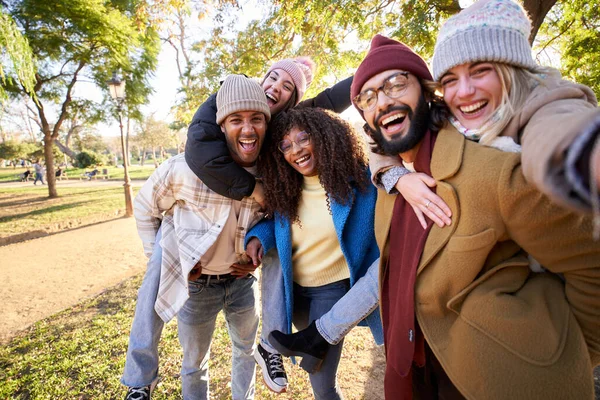 Winter selfie group excited friends smiling at camera in park on sunny day. Young multiracial millennial people gathered piggyback enjoying friendship outdoors on weekend. Relationships, leisure time.