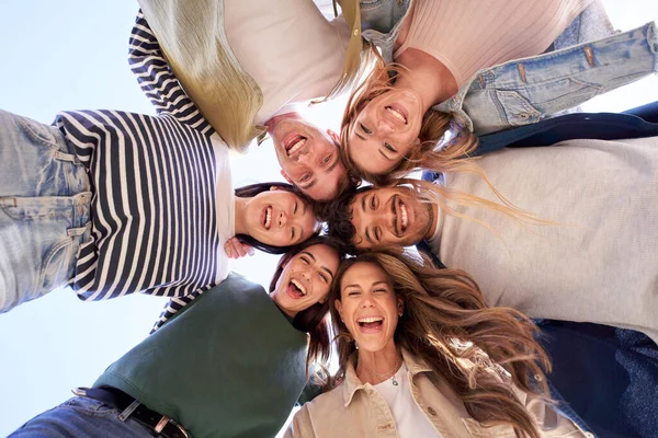 Multiracial group of young beautiful people standing in circle and smiling excited at camera. Happy diverse generation z friends having fun embracing together. Low angle view sunny portrait outdoor.